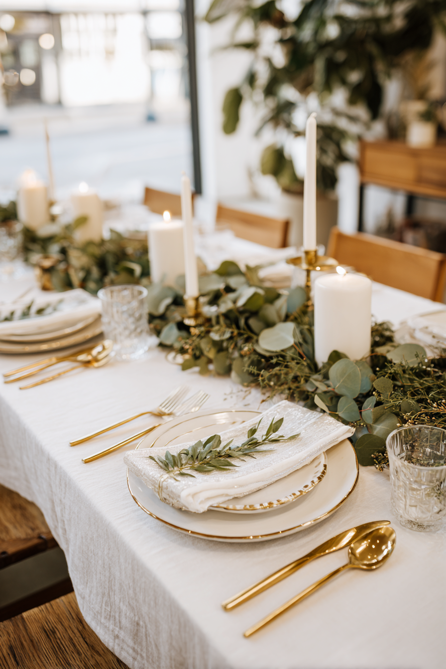 Thanksgiving table with white linens, soft gold flatware, greenery centerpiece, candles